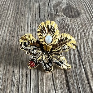 May include: A gold-toned brooch in the shape of a flower with a pearl in the centre and rhinestones around the edges. The flower has a red accent on one of the petals.