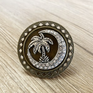 Może przedstawiać: Metalowy wisiorek w kolorze brązu z wzorem półksiężyca ozdobionym przezroczystymi cyrkoniami. Srebrna palma znajduje się w środku księżyca.