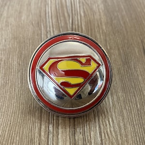 Könnte beinhalten: Ein silberfarbener Metallring mit einem roten und gelben Superman-Logo. Der Ring hat einen roten Außenrand und einen silbernen Innenrand.