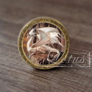 Op de afbeelding: Een ronde, messingkleurige knop met een witte draak met bruine vleugels. De draak zit op een boomstam met een bruine en oranje achtergrond.