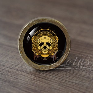 Op de afbeelding: Een ronde, antieke goudkleurige kastknop met een zwarte achtergrond en een gouden schedelontwerp. De schedel is omgeven door sierlijke gouden krulwerk.