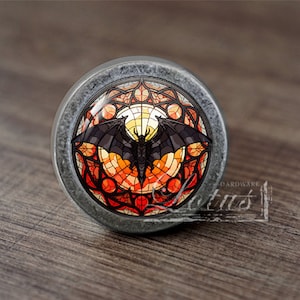May include: A round, silver-toned cabinet knob with a stained glass design featuring a black bat in a circular frame with orange, red, and yellow accents.