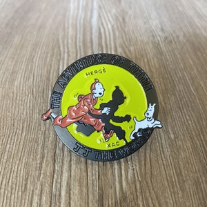 May include: A yellow and black enamel pin featuring Tintin, a cartoon character, running with his dog Snowy. The pin reads "The Adventure of Tintin" and "Hergé" in black text. The pin also reads "J.T. The World" in black text.