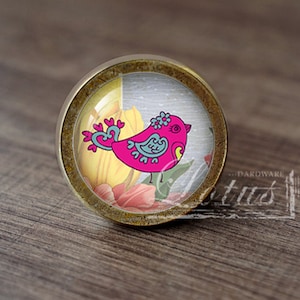 Può includere: Un pomello per armadio rotondo in ottone con un inserto in vetro che presenta un uccello rosa con accenti blu e un design floreale. L'uccello è circondato da fiori gialli e rosa.