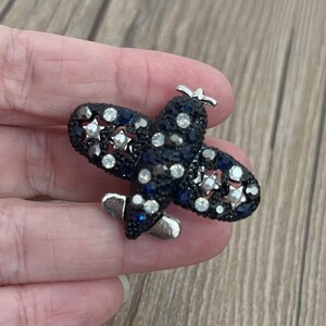 Könnte beinhalten: Eine silberne Flugzeugbrosche mit blauen und weißen Strasssteinen. Das Flugzeug hat ein Sternendesign auf den Flügeln.