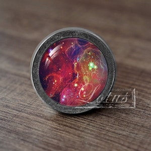 Puede incluir: Un pomo de armario redondo de color plateado con un inserto de vidrio que presenta una imagen de nebulosa colorida. La nebulosa es una mezcla de colores rojo, naranja, amarillo y azul.