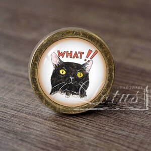 Może przedstawiać: Okrągła gałka do szafki z mosiężnym wykończeniem. Gałka przedstawia akwarelową ilustrację czarnego kota z żółtymi oczami i napisem "WHAT!!" w kolorze czerwonym. Unikalny element dekoracyjny.