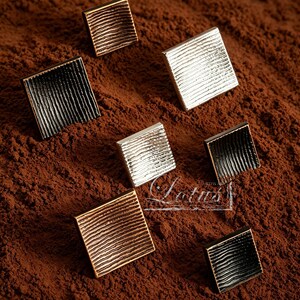 Op de afbeelding: Close-up van verschillende vierkante kastknoppen in diverse metallic afwerkingen, waaronder goud, zilver en zwart. Elke knop heeft een getextureerd, geribbeld oppervlak. De knoppen zijn gerangschikt op een bruin, getextureerd oppervlak.