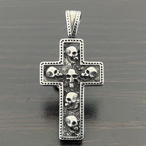 May include: A silver cross pendant with five detailed skull engravings. The cross has a beaded border.