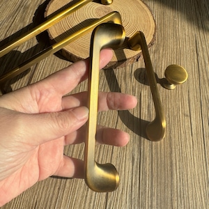 May include: Close-up of several brass cabinet pulls and a knob. The pulls have a unique, curved design with a rounded end. The hardware has a warm, antique gold finish. The items are displayed on a wooden surface.