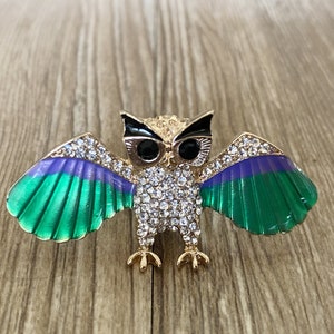 May include: A gold-tone owl brooch with green and purple enamel wings and clear rhinestone accents.