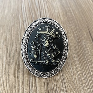 以下が含まれることがあります： 帽子をかぶって金の袋を持った白い骸骨が描かれた黒いカメオがついた銀の指輪。カメオは装飾的な銀の縁取りで囲まれています。