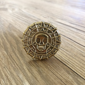 Könnte beinhalten: Eine goldfarbene Metallmünze mit einem Totenkopf- und Knochenkreuz-Design. Die Münze hat einen erhöhten Rand und eine strukturierte Oberfläche. Der Schädel ist detailliert mit einem zahnigen Grinsen und Augenhöhlen.