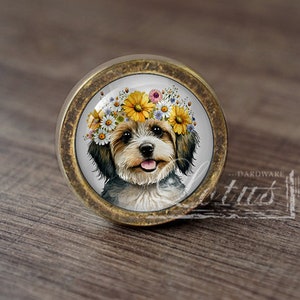 Op de afbeelding: Een ronde, messingkleurige kastknop met een witte achtergrond en een afbeelding van een hond die een bloemenkrans draagt. De hond heeft bruin en wit haar en kijkt recht in de camera.