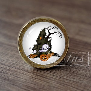 Pode incluir: Um botão de armário redondo de cor latão com um inserto de cerâmica branca que apresenta uma casa assombrada de desenho animado com fantasmas e abóboras.