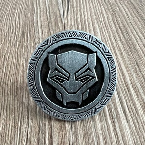 May include: A silver-colored metal coin with a black panther face design in the center. The coin has a decorative border with a repeating pattern.