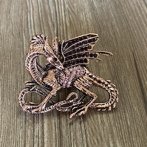 May include: A rose gold dragon brooch with pink and purple rhinestones. The dragon is curled up with its wings spread out.