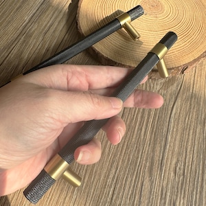 Puede incluir: Dos tiradores de armario negros con un patrón texturizado y detalles dorados. Los tiradores tienen forma cilíndrica con un tirador dorado en forma de T. Un tirador se sostiene en una mano y el otro está en el fondo.