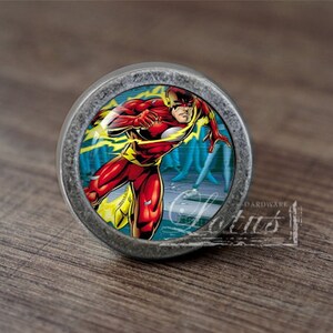 Pode incluir: Um botão de armário redondo, de cor prateada, com um inserto de vidro que apresenta uma ilustração de banda desenhada do Flash a correr através de um fundo azul e amarelo.