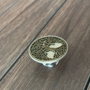 May include: A silver and gold metal ring with a tree of life design. The ring is on a wooden surface.
