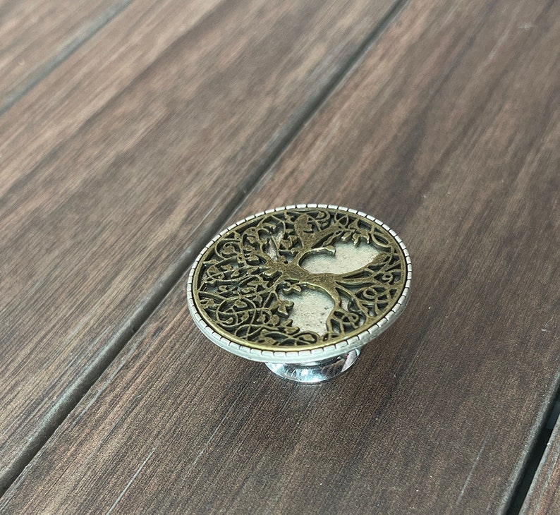May include: A silver and gold metal ring with a tree of life design. The ring is on a wooden surface.