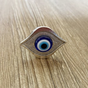 May include: A silver-coloured evil eye charm with a blue and white eye design. The charm is shaped like a teardrop and has a textured surface.