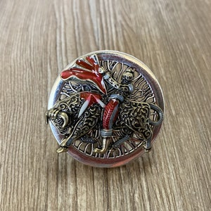 May include: A silver ring featuring a red and gold bullfighter design. The bullfighter is holding a red cape and riding a bull. The ring is set within a silver circle with a decorative design.