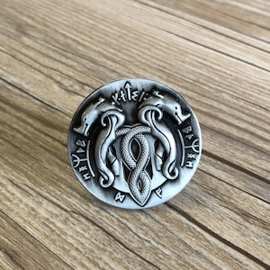 May include: A silver-toned metal pin with a design of two intertwined snakes and a symbol of a snake head with a crown. The pin is surrounded by a circle of runes.