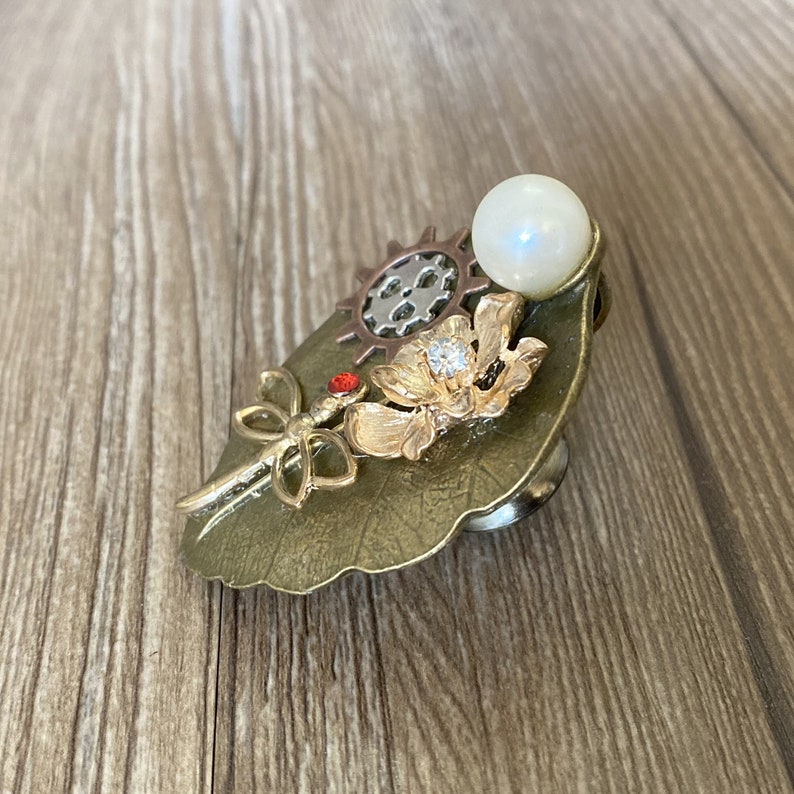 May include: A bronze leaf-shaped brooch with a pearl, a dragonfly with a red gem, a flower with a rhinestone, and a gear design.
