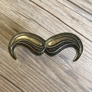 May include: A gold-toned metal mustache-shaped drawer pull. The pull has a detailed design with multiple curves and lines.
