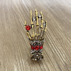 K&ouml;nnte beinhalten: Goldene Skeletthand mit roten und klaren Strasssteinen. Die Hand h&auml;lt einen Strau&szlig; aus roten und klaren Strasssteinen.