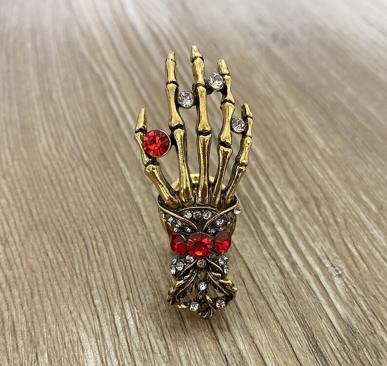 K&ouml;nnte beinhalten: Goldene Skeletthand mit roten und klaren Strasssteinen. Die Hand h&auml;lt einen Strau&szlig; aus roten und klaren Strasssteinen.