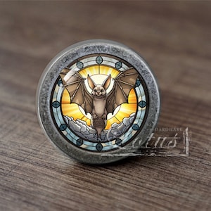 May include: A round, silver-coloured cabinet knob with a stained glass design featuring a bat flying in front of a sun and clouds. The knob has a raised edge and a smooth surface.