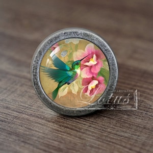 Puede incluir: Un pomo de plata con un inserto de vidrio redondo que presenta un colibrí en vuelo con flores rosas.