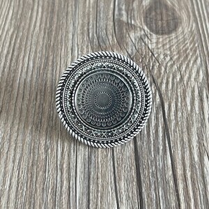 以下が含まれることがあります： 円形のデザインで、同心円のパターンとロープのような縁取りが施された銀の指輪です。