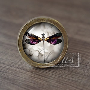 May include: A round, brass-coloured cabinet knob with a glass insert featuring a purple and brown dragonfly on a white background.