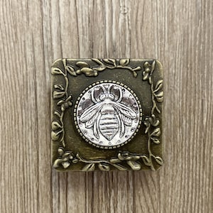 May include: A square, antique gold-toned metal box with a floral design. The box has a circular, silver-toned bee design in the center.