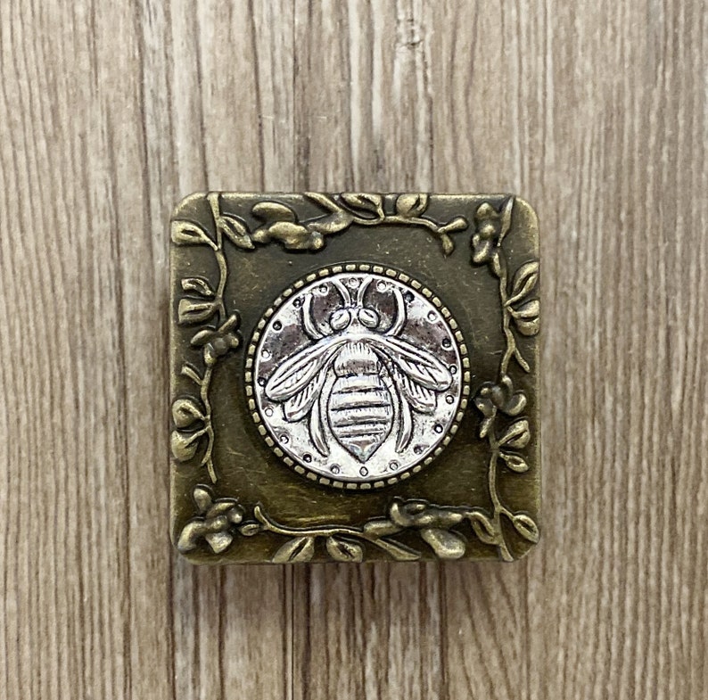 May include: A square, antique gold-toned metal box with a floral design. The box has a circular, silver-toned bee design in the center.