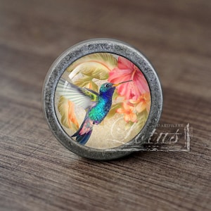 Puede incluir: Un pomo de armario redondo de color plateado con un inserto de vidrio que presenta un colibrí colorido en vuelo sobre flores rosas y amarillas.