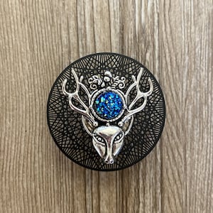 Op de afbeelding: Een zilveren hertenkop charme met een blauw glitter middenstuk, op een zwarte ronde achtergrond met een decoratief patroon.