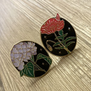 May include: Two round enamel pins with black backgrounds and gold outlines. One pin features a red poppy flower with green leaves and stems. The other pin features a purple hydrangea flower with green leaves and stems.