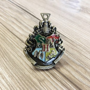 May include: A gold-colored metal Hogwarts house crest with a green, red, yellow, and blue enamel design. The crest features a lion, a badger, a snake, and an eagle. The words "Draco Dormiens Nunquam Titillandus" are inscribed around the crest.