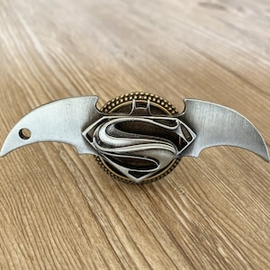 May include: A silver and gold metal Batman batarang with a Superman logo in the center. The batarang is on a wooden surface.