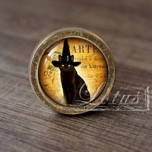 Może przedstawiać: Okrągła gałka do szafki w stylu antycznym z czarnym kotem w kapeluszu czarownicy. Kot jest umieszczony na tle w stylu vintage z widocznym słowem "ARTE". Gałka ma brązowe wykończenie.