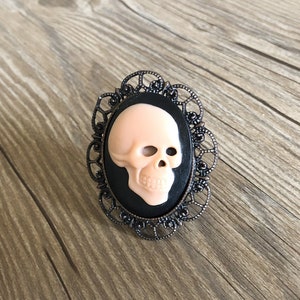 May include: A black cameo ring with a detailed filigree design. The cameo features a white skull with a black background.