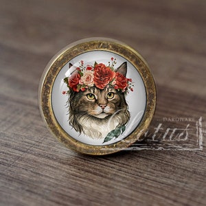 May include: A round, brass-coloured cabinet knob with a glass insert featuring a brown and white cat wearing a red and white flower crown.