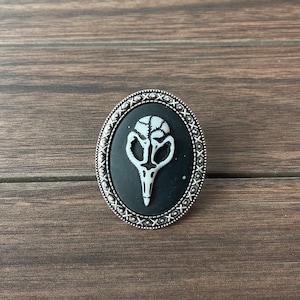 Op de afbeelding: Een zilveren ovale broche met een zwarte achtergrond en een wit vogelskul design.