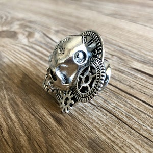 May include: A silver skull ring with intricate gear details. The skull is detailed with a stitched design on the forehead.