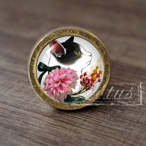 May include: Circular cabinet knob with a cat design. The knob shows a black and white cat profile, pink dahlia, and red and yellow flowers. It has a brass-coloured metal edge and a green bow.