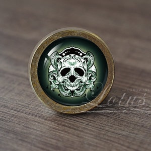 Op de afbeelding: Een ronde kastknop met een schedelontwerp. De knop heeft een messingkleurige metalen basis en een glazen inzetstuk met een witte schedelafbeelding op een donkergroene achtergrond. De schedel is omgeven door sierlijke, wervelende details.